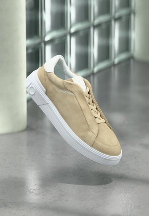 Baskets en daim beige avec semelle blanche et lacets, de la marque "Valentino", flottant au-dessus d'un sol en béton poli près d'un mur en verre.