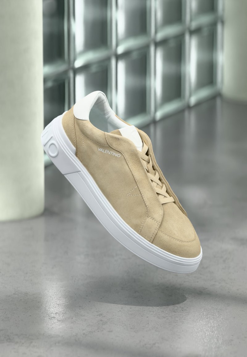 Baskets en daim beige avec semelle blanche et lacets, de la marque "Valentino", flottant au-dessus d'un sol en béton poli près d'un mur en verre.