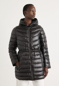 Doudoune noire avec capuche, présentant un design matelassé, une taille avec cordon de serrage, des poches latérales et une texture brillante.