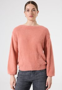Flauschiger, korallenroter Pullover mit weitem Ausschnitt und langen Ärmeln, mit lockerer Passform und weichem Material, kombiniert mit dunkelgrauen Jeans.