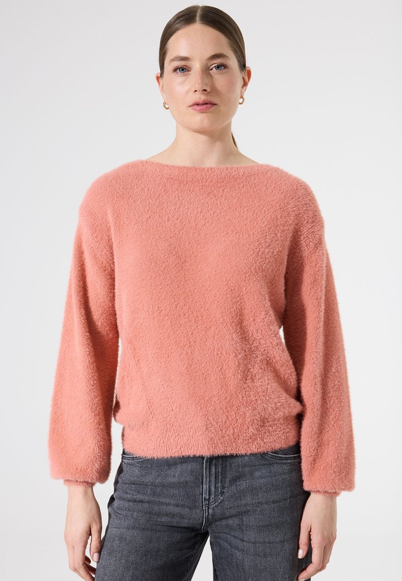 Flauschiger, korallenroter Pullover mit weitem Ausschnitt und langen Ärmeln, mit lockerer Passform und weichem Material, kombiniert mit dunkelgrauen Jeans.