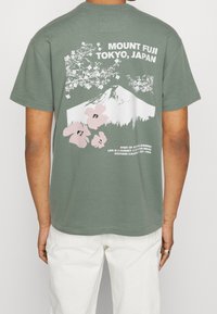 Olive green cotton t-shirt featuring a graphic of Mount Fuji, pink blossoms, and white lettering on the back. Short sleeves and relaxed fit.