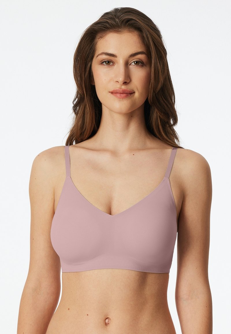 Woman wearing a light mauve seamless, wireless bra with thin adjustable straps, standing against a plain white background.