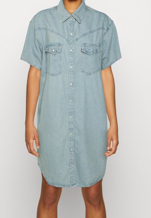 Robe chemise en jean bleu clair à manches courtes avec boutons-pression à l'avant et deux poches à rabat sur la poitrine, portée par une personne debout.