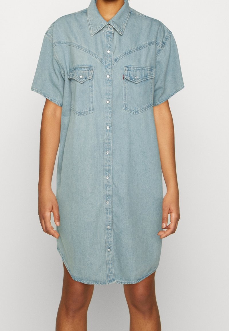 Robe chemise en jean bleu clair à manches courtes avec boutons-pression à l'avant et deux poches à rabat sur la poitrine, portée par une personne debout.