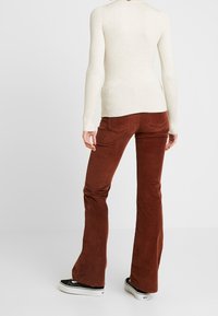 Personne portant un pantalon flare en velours côtelé marron, un haut à manches longues côtelé crème et des baskets noires, debout devant un fond uni.