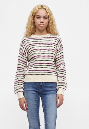 Ung kvinde med krøllet blondt hår iført en hvid strikket sweater med pink og sorte vandrette striber og blå jeans, stående mod en hvid baggrund.