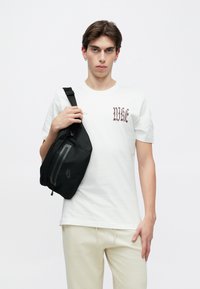 Czarna torba na ramię o gładkiej fakturze, z przednią kieszenią na zamek i detalem z logo, noszona na białej koszulce i jasnych spodniach.