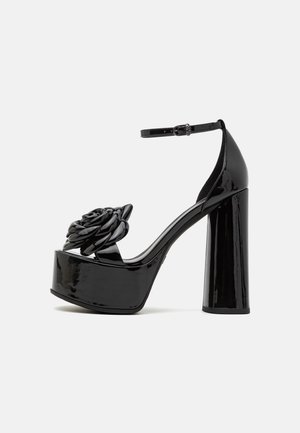 Czarne sandały na platformie z lakierowanej skóry, o masywnym obcasie, zdobione dekoracyjnym kwiatem z przodu oraz regulowanym paskiem wokół kostki.