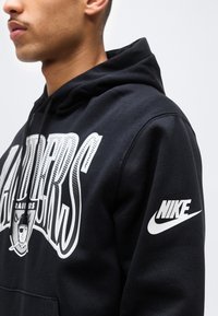 Νεαρός που φοράει μαύρο φούτερ με κουκούλα με το λευκό λογότυπο των Raiders στο στήθος και το λευκό λογότυπο της Nike στο μανίκι, κοιτάζοντας αριστερά σε απλό φόντο.