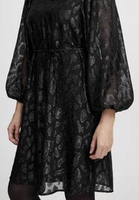 Rochie neagră cu un overlay transparent având un model de imprimeu leopard metalic. Are mâneci bufante și talie strânsă cu un șnur.