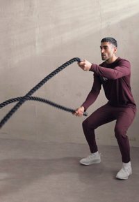 Sorte battle ropes holdes af en person i bordeaux træningstøj og hvide sneakers, der udfører en øvelse i et minimalisk indendørs rum.