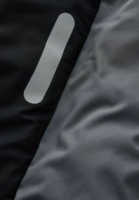 Detailný záber čierneho a šedého textilu s vertikálnym reflexným prúžkom na čiernej časti, ktorý zobrazuje textúru a detaily šitia.