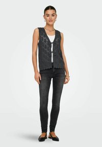 Gilet grigio lavorato a maglia con un motivo geometrico, aperto davanti e con chiusura a nodo, indossato sopra una maglietta bianca e abbinato a jeans skinny scuri.