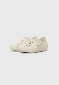 Nike-träskor i ljusbeige med ett vågigt mönster, flera ventilationshål och en reflekterande silverlogotyp på sidan. Strukturerad gummisula.