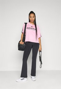 Kvinne iført rosa Adidas treningsdrakt, svarte flagrende tights, hvite Adidas sneakers, bærende på svart treningspose og holder en hoppetau.