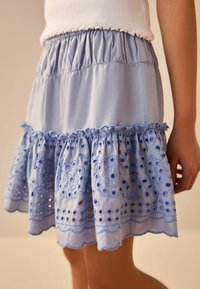 Jupe en coton bleu clair avec taille froncée, broderie à œillets, ourlet festonné et couches volantées. Texture subtile dans le tissu et le design.