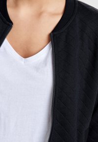 Närbild av en person som bär en vit V-ringad t-shirt och en ofodrad svart quiltad jacka med ribbad krage.