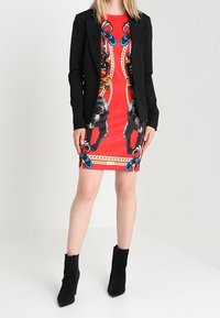 Veste blazer noire cintrée portée sur une robe rouge ornée de motifs audacieux d'animaux, de chaînes et d'accents bleus. Portée avec des bottines noires.