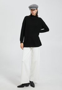 Schwarzer gerippter Rollkragenpullover mit langen Ärmeln, kombiniert mit weißen weit geschnittenen Hosen und schwarzen spitzen Ankle Boots, accessoriert mit einer schwarz-weißen karierten Mütze.