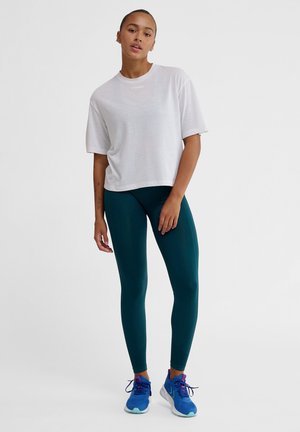White short-sleeve t-shirt, teal leggings, and blue athletic shoes. The t-shirt features a subtle logo. The fabric appears soft and comfortable.