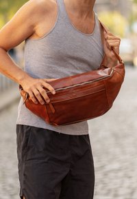 Brown leather crossbody bag with a zippered pocket, rounded shape, smooth texture, and a strap being held by a person wearing a grey tank top.