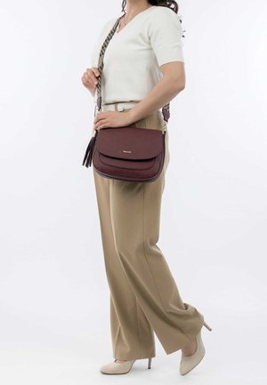 Mujer con pantalones beige y camiseta blanca sosteniendo un bolso cruzado color granate con una correa ancha con estampado y detalle de borlas.