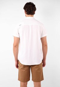 Chemise blanche à manches courtes avec un col boutonné, texture lisse et ourlet arrondi. Associée à un short beige, elle présente un style décontracté.