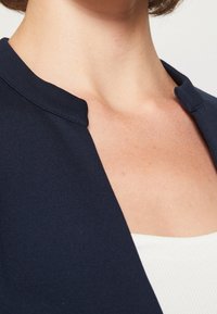 Granatowa marynarka o gładkim materiale, z wyraźnym kołnierzem i otwartym przodem, podkreślająca prosty, minimalistyczny design.