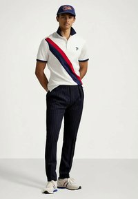 Νεαρός άνδρας που φοράει άσπρο πόλο με κόκκινες και ναυτικές διαγώνιες ρίγες, ναυτικά παντελόνια με ρίγες, άσπρα αθλητικά παπούτσια και ναυτικό καπέλο με κόκκινο λογότυπο.