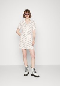 Vestido de manga curta, com decote em V, em tecido leve com um padrão floral apresentando pequenas flores vermelhas. Combinado com botas brancas de atacadores.