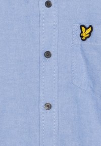 Camisa de botones azul claro con un pequeño logo de águila bordado en amarillo sobre el bolsillo izquierdo del pecho.