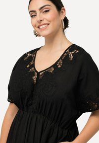 Vestido negro con mangas cortas, que presenta intrincados bordados de encaje negro en el escote y una cintura fruncida. Textura de tela suave.