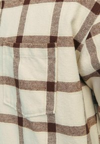 Chemise en flanelle à carreaux aux teintes beige clair et marron, avec une surface texturée et une poche sur la poitrine à gauche.
