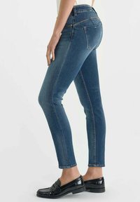 Jeans skinny en denim bleu foncé, avec une taille haute, cinq poches, des coutures discrètes et une texture lisse. Stylés avec des mocassins noirs.