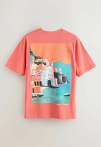 T-shirt corail avec une illustration colorée des falaises de la côte d'Amalfi, des bâtiments, de la mer et d'un voilier sous un ciel orange.