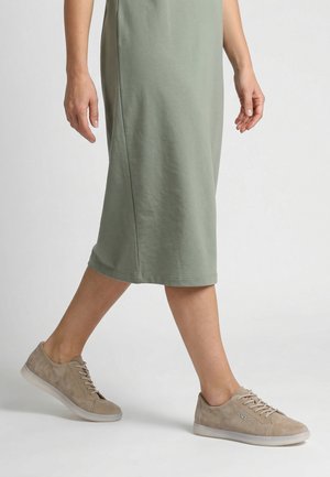 Beine in beigen Sneakers gehen neben einer Person, die einen knielangen salbeigrünen Rock trägt, vor einfarbigem Hintergrund.