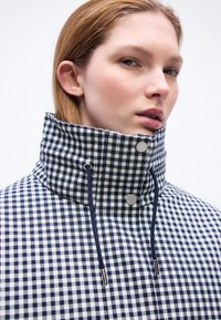 Γυναίκα με ανοιχτόχρωμο δέρμα και ανοιχτό καφέ μαλλιά, που φοράει μπλε και άσπρο καρό μπουφάν με ψηλό γιακά, κορδόνια και μεταλλικά κουμπιά.