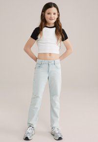 Camisa blanca acanalada recortada con mangas negras, combinada con unos vaqueros desgastados de color azul claro. El modelo lleva zapatillas grises. Un atuendo simple y casual.