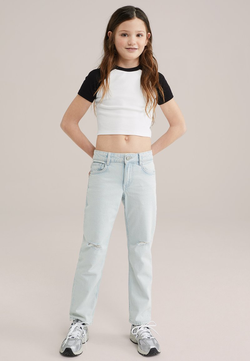 Camisa blanca acanalada recortada con mangas negras, combinada con unos vaqueros desgastados de color azul claro. El modelo lleva zapatillas grises. Un atuendo simple y casual.