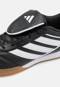 Black leather athletic shoe featuring three white stripes, lace-up design, perforated accents, and a gum rubber sole.