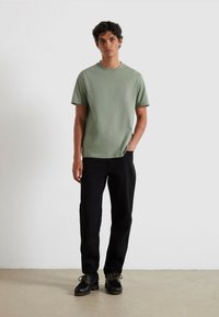 T-shirt da uomo a maniche corte in cotone verde tenue, con un logo sottile. Abbinato a pantaloni neri a gamba dritta e scarpe nere con lacci.