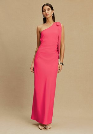 Žena v ružových šatách dĺžky po zem s jedným ramenom a mašľou, doplnených kruhovými náušnicami, náramkom a zdobenými podpätkami.