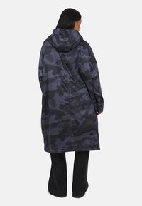 Donna che indossa un lungo cappotto mimetico blu scuro e nero, ampio, con cappuccio, in piedi di spalle, con pantaloni neri e scarpe scure.