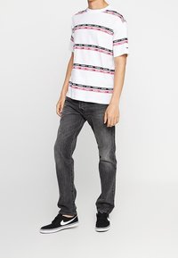 Camiseta blanca de manga corta con rayas horizontales negras y rosas de marca, combinada con jeans gris oscuro y zapatillas negras con suelas blancas.