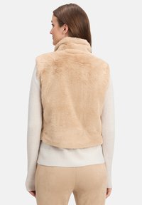 Beżowa kamizelka z sztucznego futra z wysokim kołnierzem, krótki krój i miękka tekstura. Dopasowana do jasnego swetra i pasujących spodni w stonowanej palecie. 
