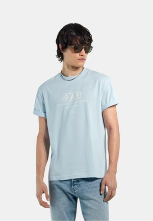 Giovane uomo con maglietta azzurra a maniche arrotolate con logo, jeans blu, occhiali da sole e catena al collo, che guarda di lato.