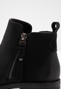 Stivaletto in suede nero con chiusura a zip, nappina decorativa e suola piatta. Presenta una punta arrotondata e una texture liscia.