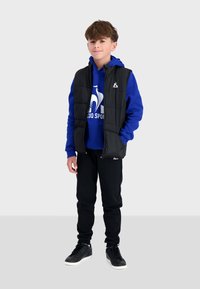 Gilet rembourré noir sur un sweat à capuche bleu avec logo blanc, joggeurs noirs et sneakers noirs avec semelles blanches, se tenant sur un fond neutre.