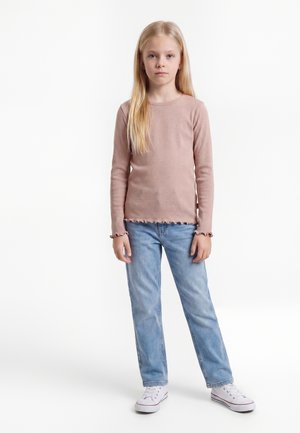 Jeune fille aux longs cheveux blonds portant un t-shirt rose à manches longues, un jean bleu clair et des baskets blanches, debout sur un fond blanc.